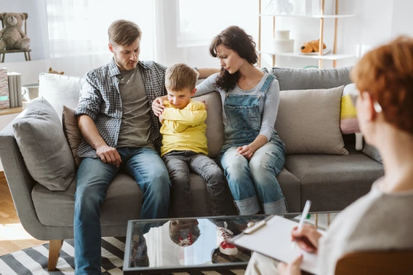 A family of three—father, mother, and child—sitting closely together on a couch, looking at someone or something out of frame.