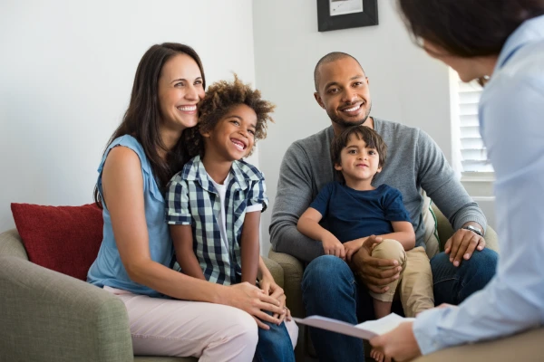 A diverse family, including a mother, father, and two children, sitting together and engaging in a conversation with a person whose back is visible in the foreground.