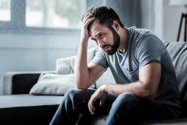 A man sitting on a couch, holding his head with his hand, appearing frustrated or distressed.