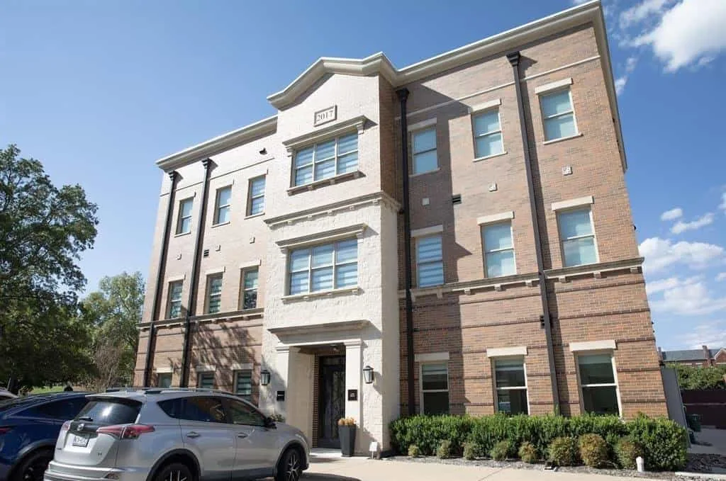 The exterior of a modern brick building with multiple windows, cars parked outside, and neatly trimmed bushes.