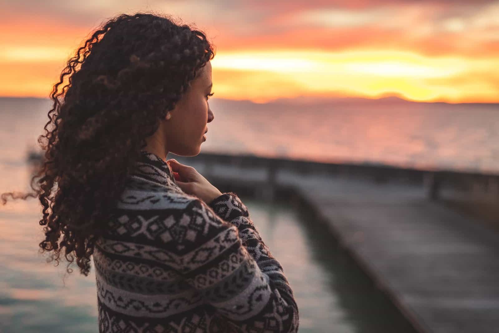 A woman wrapped in a cozy sweater, standing on a beach or boardwalk, looking thoughtfully at a vibrant sunset.