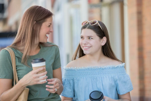 Two women chatting and smiling at each other while holding coffee cups, possibly in a city street.