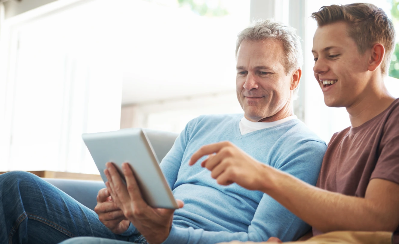 An older man smiling as he uses a tablet, with a younger man beside him, both appearing happy and engaged.