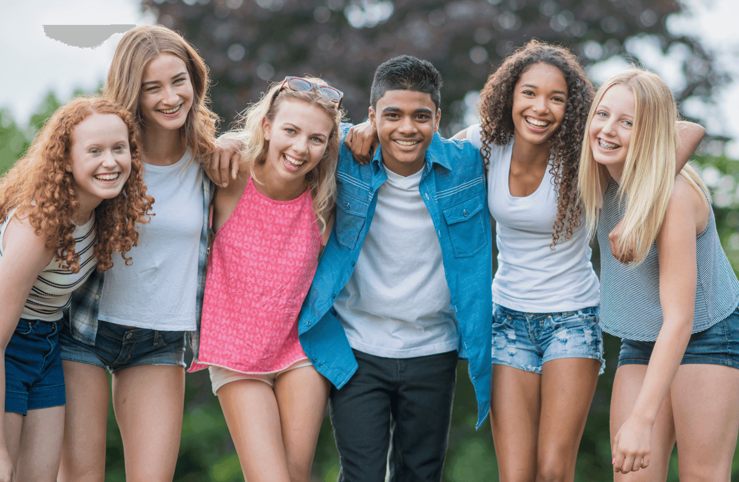 A group of six smiling teenagers, five girls and one boy, stand closely together outdoors with their arms around each other, posing happily for the camera. Trees and greenery are visible in the background.