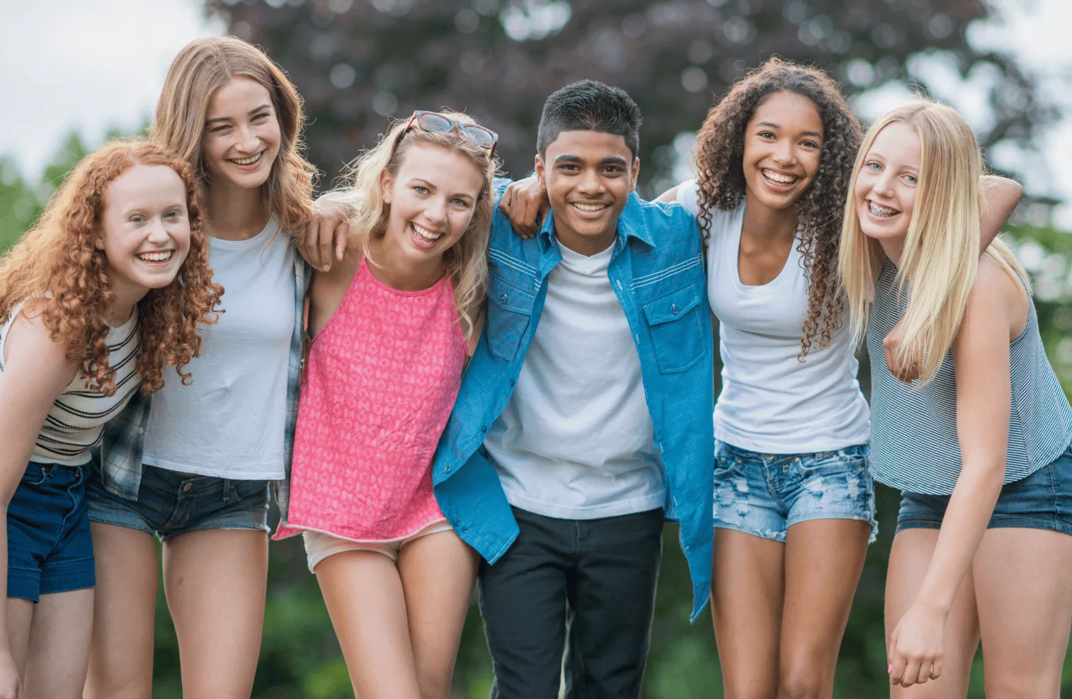 A group of six smiling teenagers, five girls and one boy, stand closely together outdoors with their arms around each other, posing happily for the camera. Trees and greenery are visible in the background.