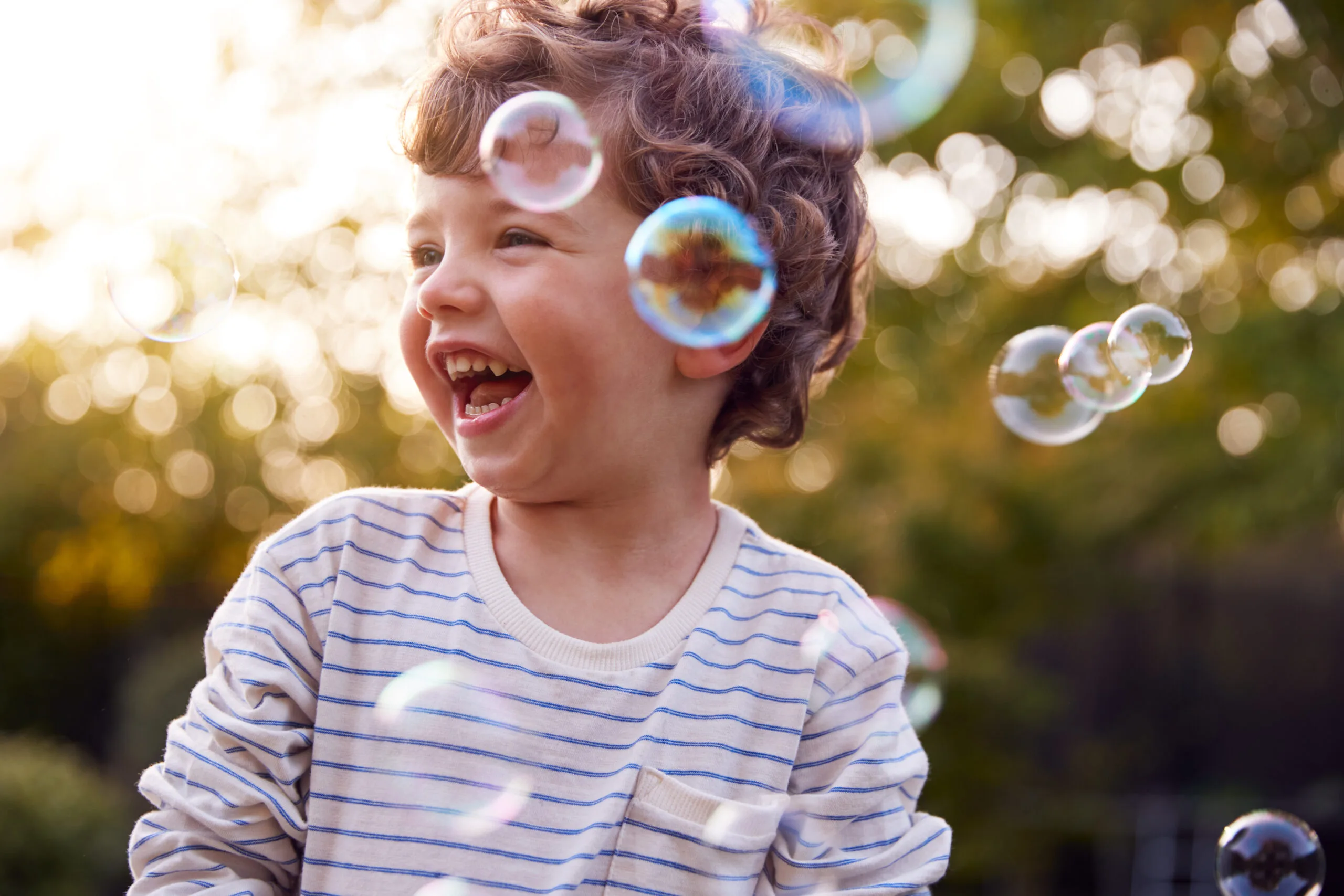 A young child with curly hair and a striped shirt laughs joyfully outdoors, surrounded by floating soap bubbles, with sunlight and greenery in the background.