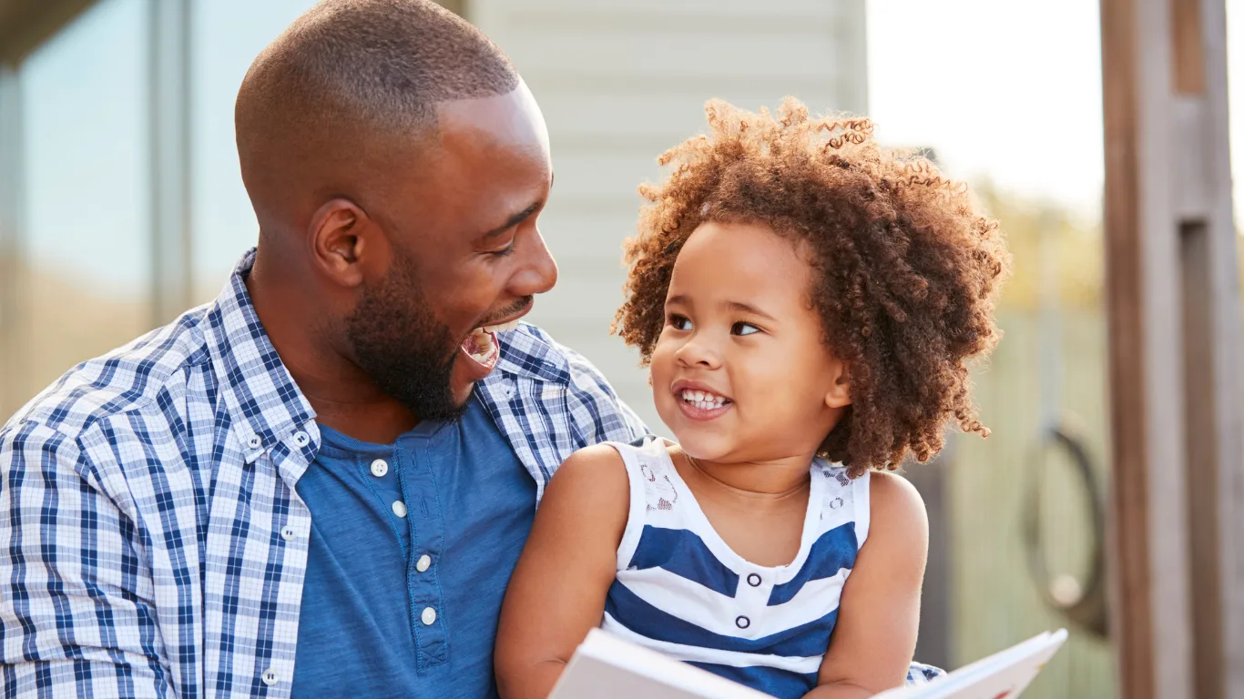 A smiling man in a plaid shirt and a young child in a striped dress sit close together outdoors, looking at each other and laughing while holding an open book.