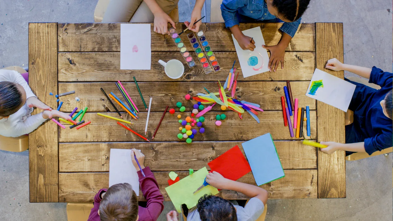 Top view of a group of children sitting around a wooden table, drawing and crafting with colored paper, markers, paint, pom-poms, and craft sticks. Their hands are busy creating art projects.