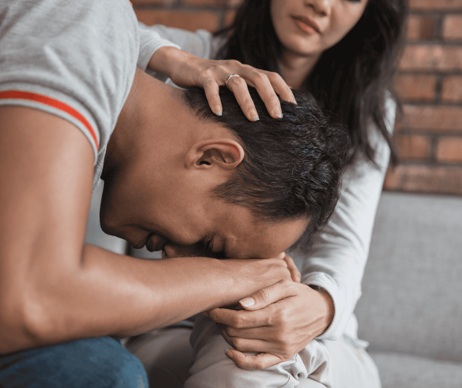 A distressed man sits with his head in his hands on a woman’s lap while she gently comforts him, placing her hand on his head and shoulder. They are sitting on a couch against a brick wall.