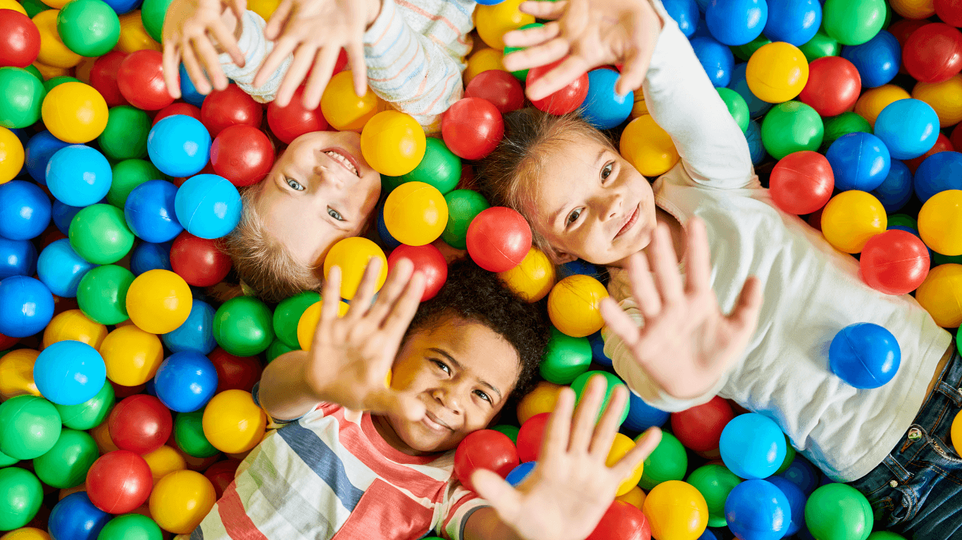 Three smiling children lying in a colorful ball pit, reaching their hands up toward the camera, surrounded by red, yellow, blue, and green plastic balls.