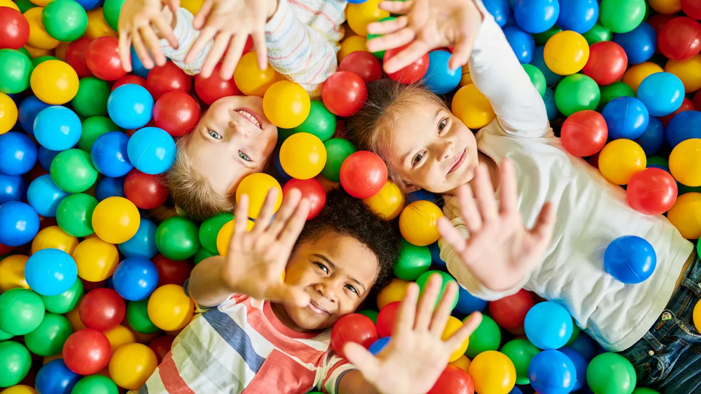 Three smiling children lying in a colorful ball pit, reaching their hands up toward the camera, surrounded by red, yellow, blue, and green plastic balls.