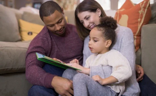 A man and woman sit closely together on a couch, smiling as they read a book with a young girl sitting on the womans lap. The family appears engaged and happy in their cozy living room.