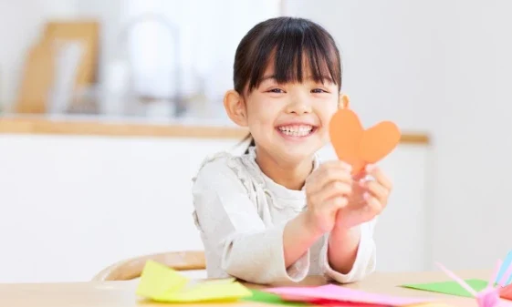 A smiling young girl sits at a table holding up an orange paper heart. Colorful sheets of paper are scattered in front of her, suggesting she is doing a craft activity.