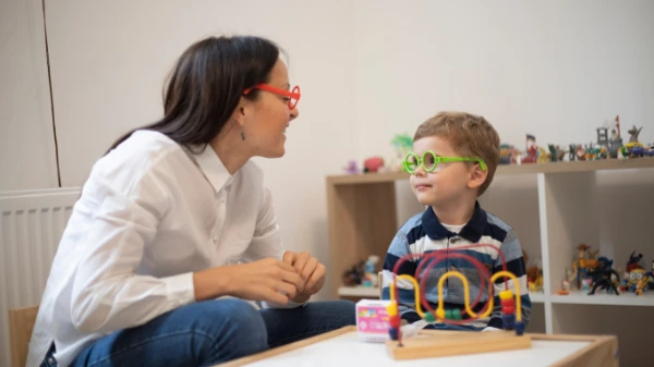 A woman and a young boy wearing colorful glasses sit together, smiling and talking. The boy is playing with a bead maze toy on a table, and shelves with toys are visible in the background.