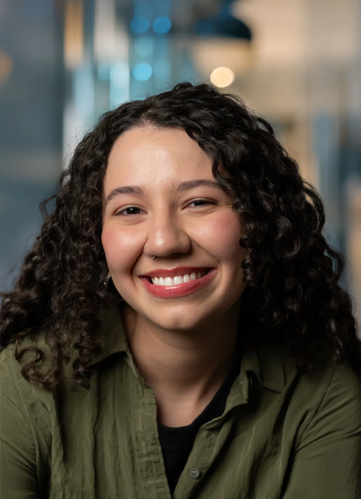 A young woman with curly dark hair, wearing a green shirt, smiles warmly at the camera. The background is softly blurred with blue and beige tones.