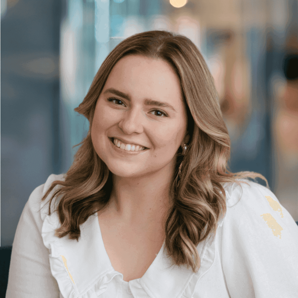 A young woman with wavy light brown hair, wearing a white blouse with ruffled details, sits smiling in a bright indoor setting with blurred blue and beige background.