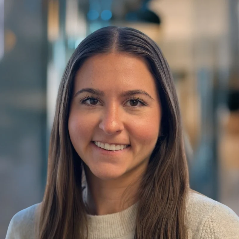 A young woman with long brown hair, wearing a light gray sweater, smiles at the camera in a softly lit indoor setting with a blurred background.