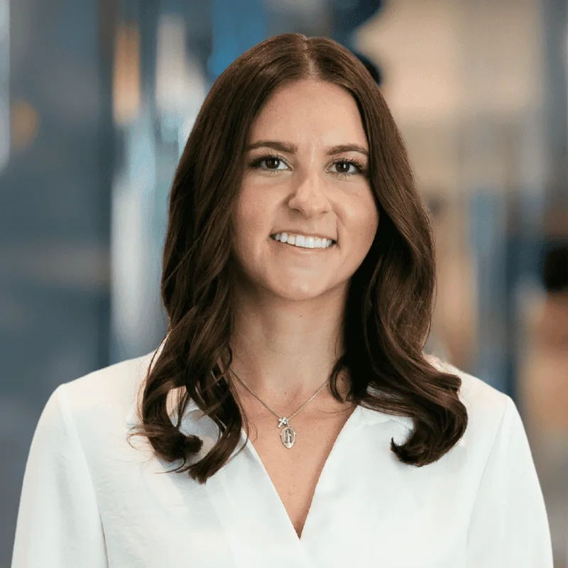 A woman with long brown hair wearing a white blouse and a necklace smiles at the camera. The background is softly blurred, suggesting an indoor, professional setting.