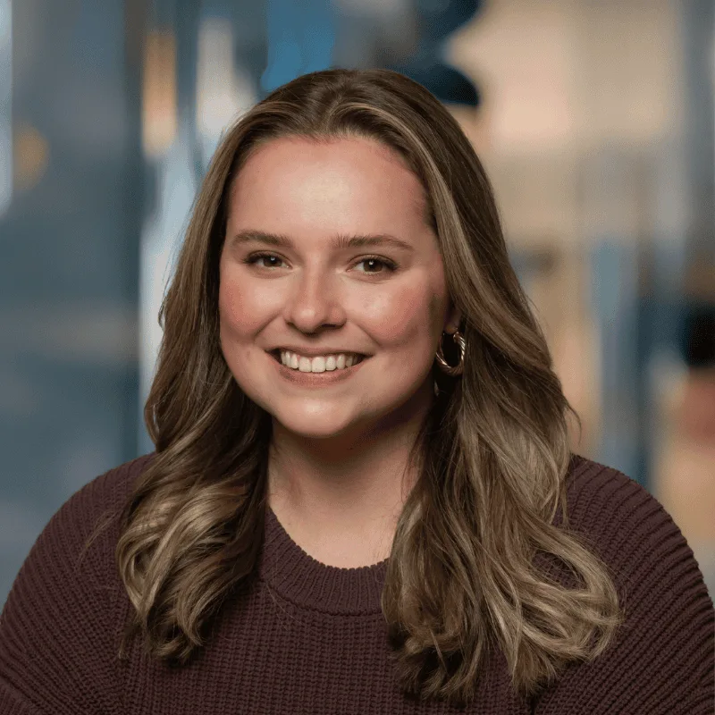 A woman with long, wavy, light brown hair, wearing a brown sweater and gold hoop earrings, smiles at the camera in an office setting with a blurred background.