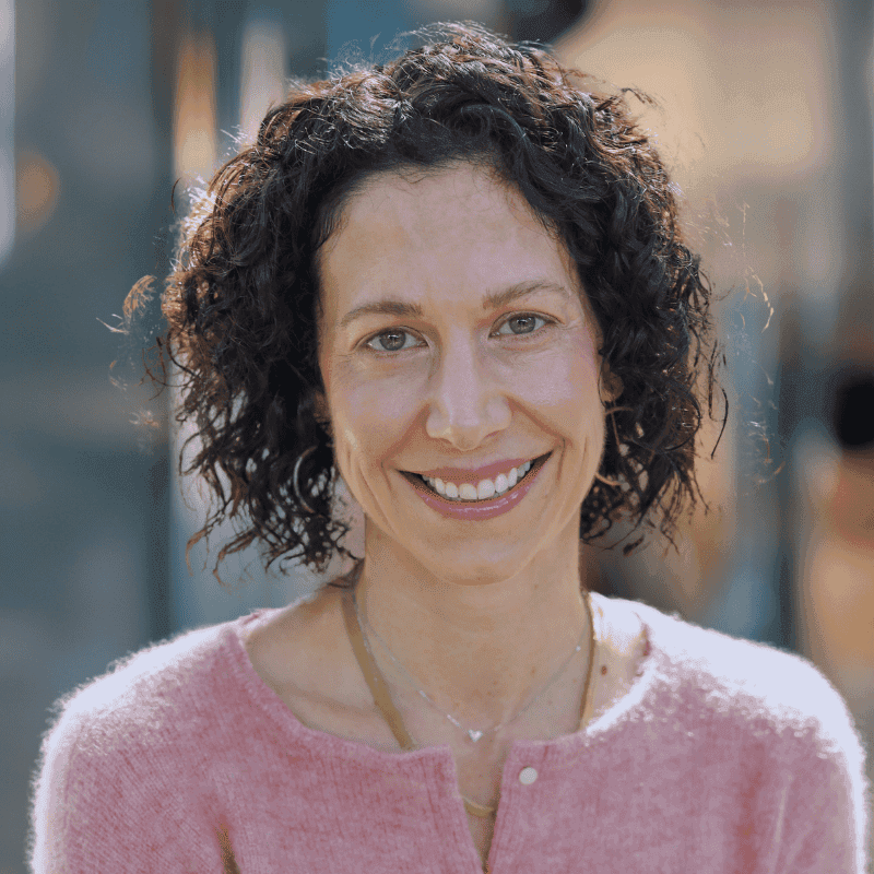 A woman with curly dark hair and fair skin smiles at the camera. She is wearing a light pink sweater and gold jewelry, and is outdoors with blurred greenery and buildings in the background.