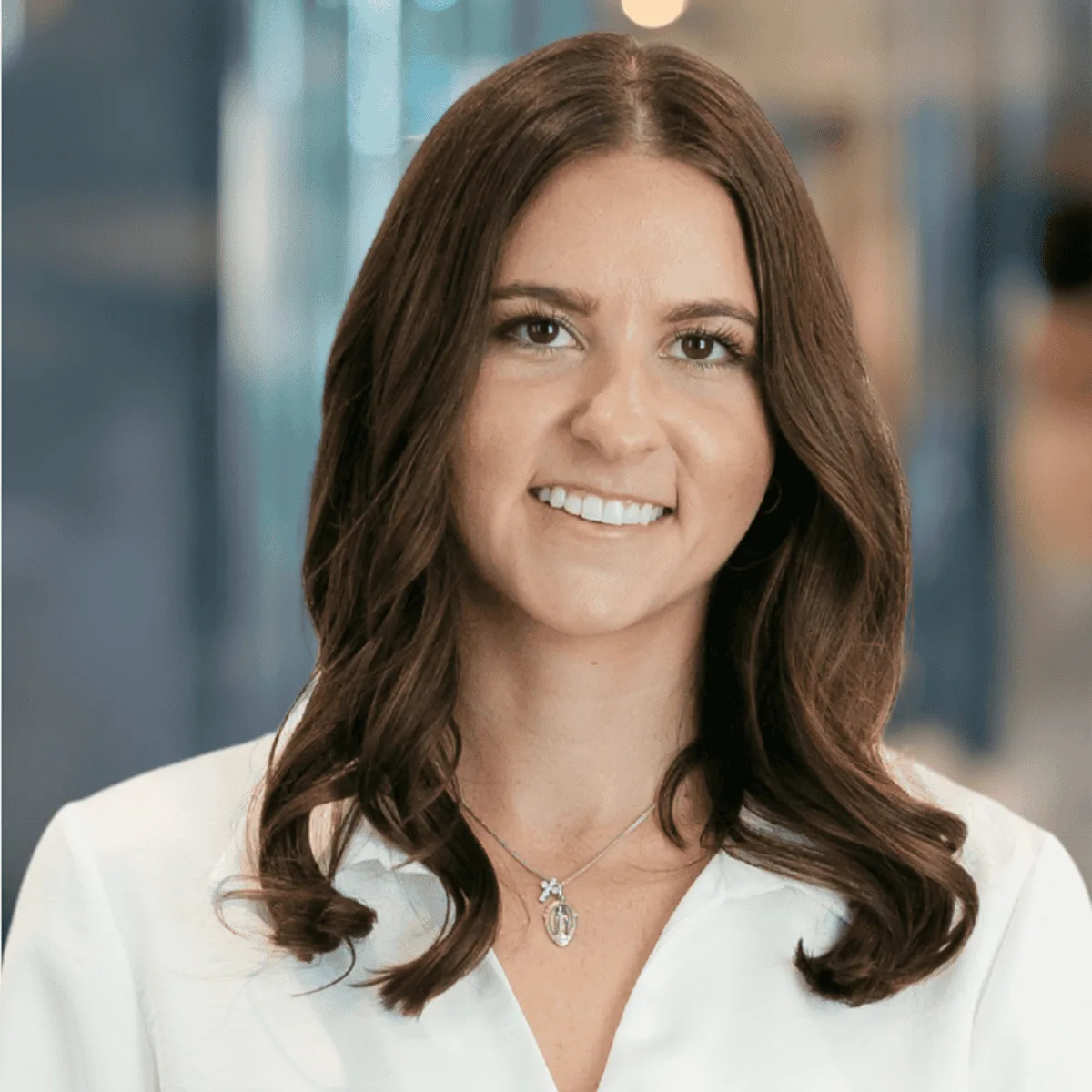 A woman with long brown hair and a white blouse smiles at the camera. She is wearing a silver necklace and is standing in a modern, softly blurred indoor setting.
