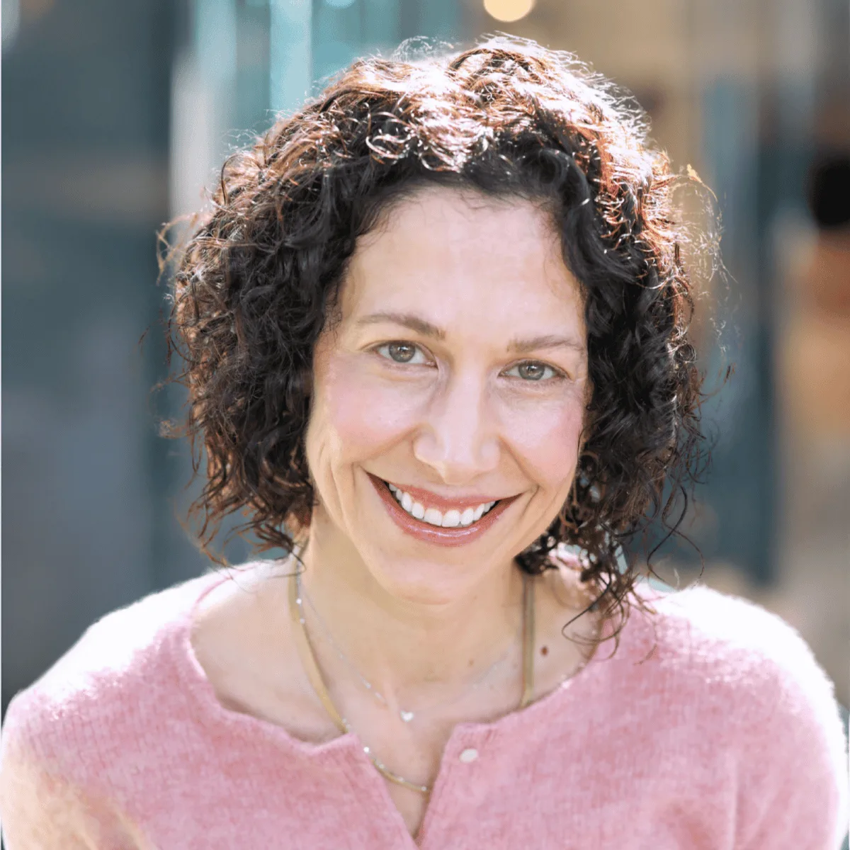 A woman with curly dark hair, wearing a light pink sweater and layered necklaces, smiles warmly at the camera in a brightly lit, softly blurred indoor setting.