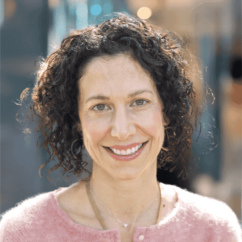 A woman with curly dark hair and light skin smiles at the camera. She is wearing a pink sweater and a gold necklace, standing in an outdoor setting with soft, blurred lights in the background.