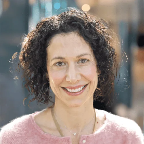 A woman with curly dark hair and light skin smiles at the camera. She is wearing a pink sweater and a gold necklace, standing in an outdoor setting with soft, blurred lights in the background.