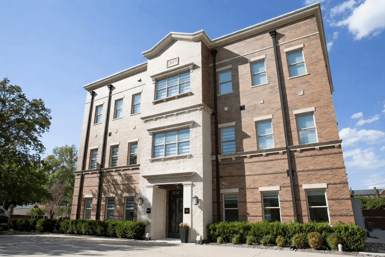 Three-story brick building with white stone accents, rectangular windows, and shrubs lining the front. The sky is clear and blue, and sunlight brightens the facade.