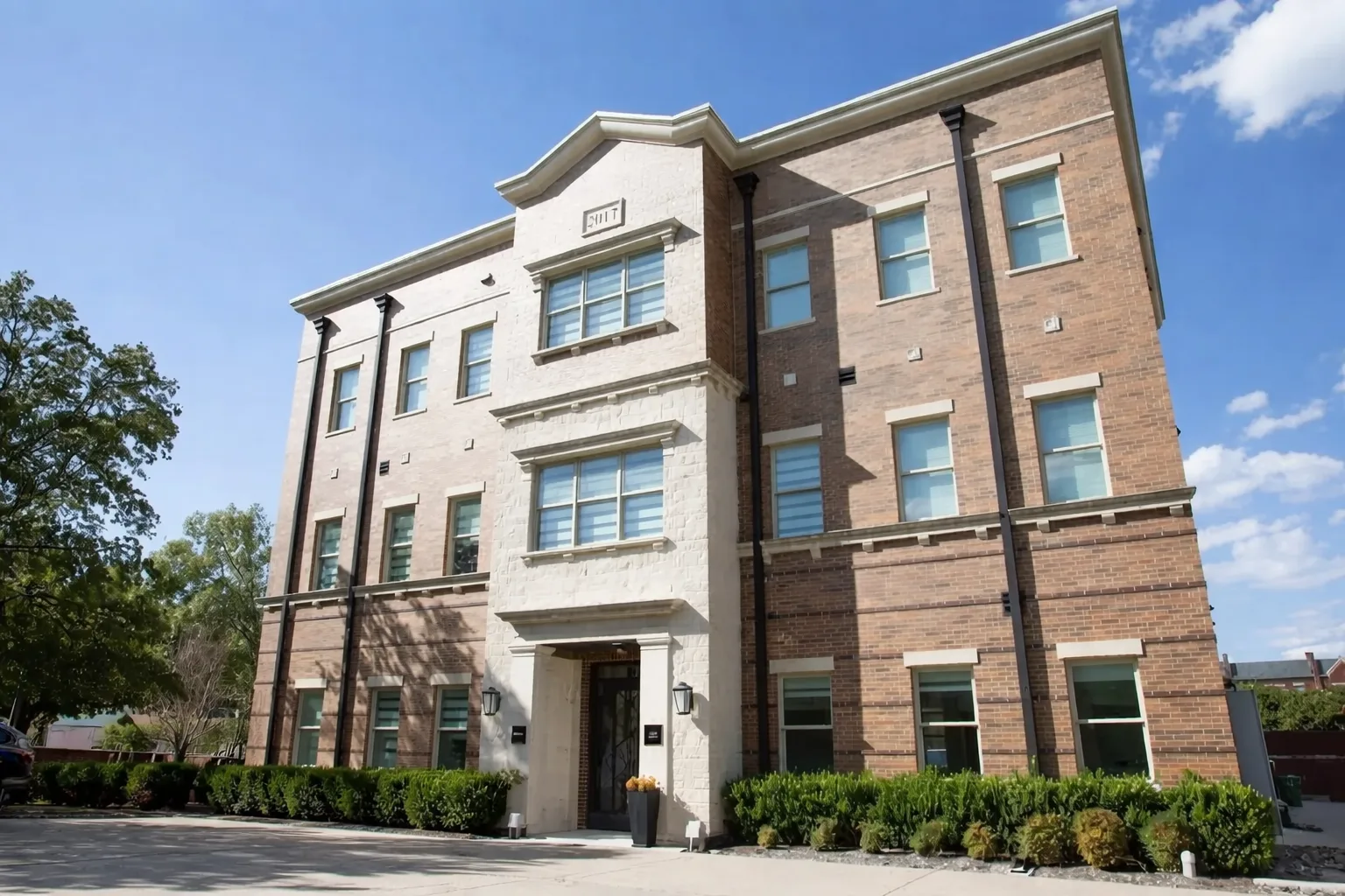 Three-story brick building with white stone accents, rectangular windows, and shrubs lining the front. The sky is clear and blue, and sunlight brightens the facade.