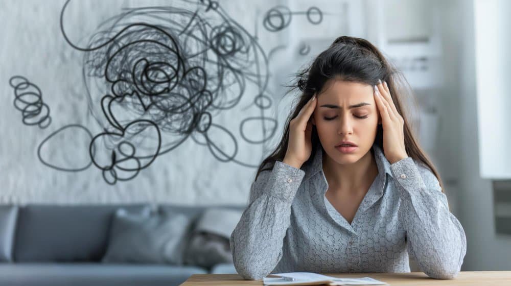 A woman sits at a table holding her head with both hands, eyes closed, looking stressed. Scribbled lines above her head symbolize confusion or anxiety—often signs that adhd counseling could provide valuable support. The background is a modern, light-colored room.