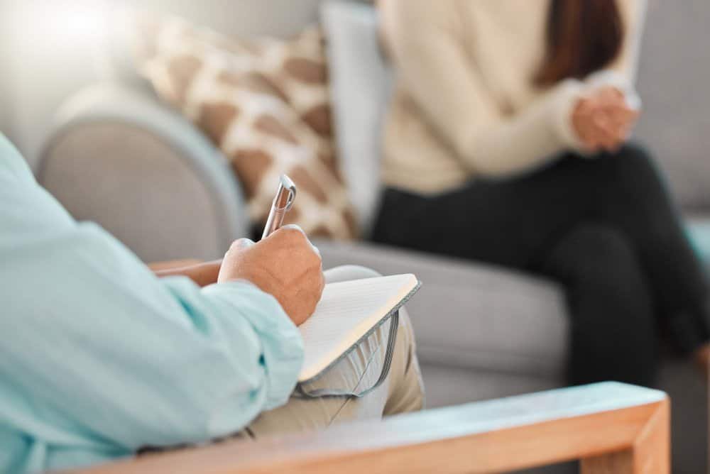A person with a notepad and pen listens and takes notes while another sits across from them on a couch, suggesting an ADHD counseling session. Both individuals are partially out of frame.