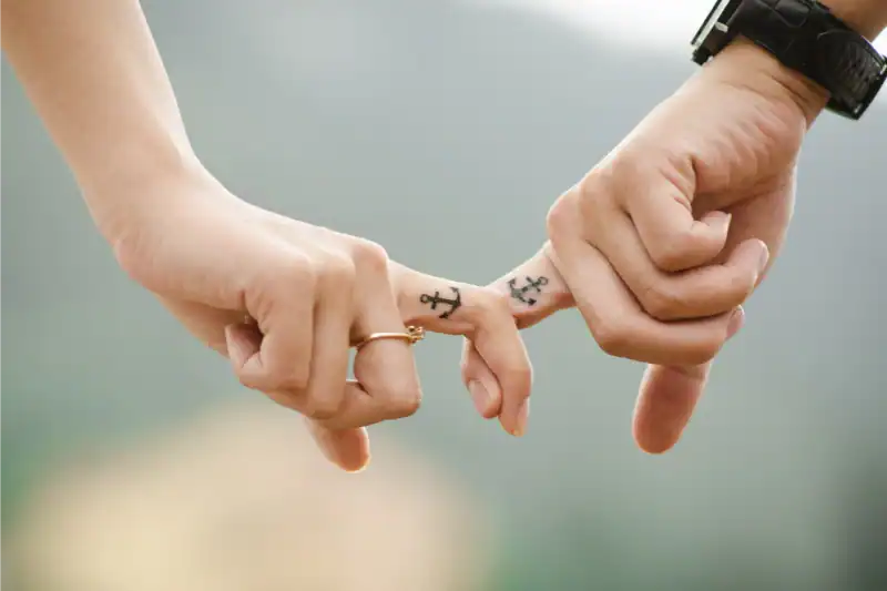 Two hands linking pinky fingers, each with a small anchor tattoo on the side of the finger, symbolizing connection and commitment. One hand has a ring; the other wears a watch. The background is softly blurred.