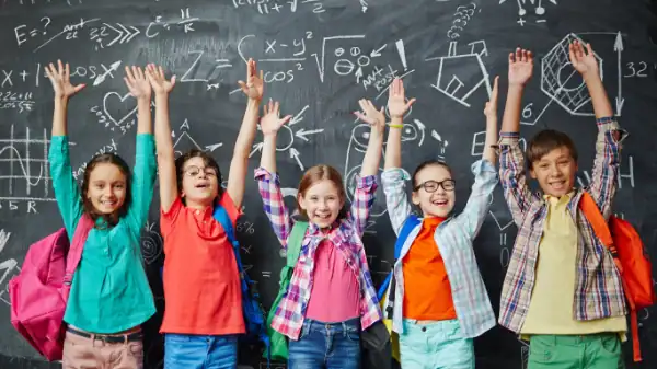 Five cheerful children with backpacks stand in front of a chalkboard covered in math and science drawings, smiling and raising their hands enthusiastically—showcasing the positive impact of ADHD counseling on classroom confidence and participation.