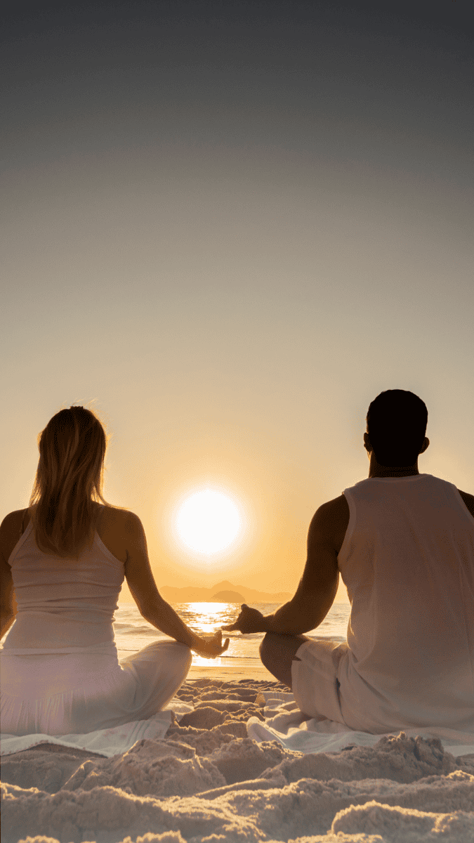 Two people sit cross-legged on the beach at sunrise or sunset, facing the ocean and holding hands, with calm waves and a glowing sun on the horizon. They appear to be meditating or enjoying a peaceful moment together.