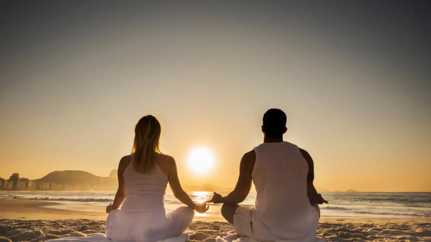 A man and a woman sit cross-legged on a sandy beach, facing the ocean and meditating at sunrise or sunset. Both wear white clothing, and the sky is glowing with warm colors.