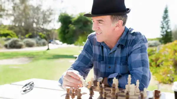A smiling man in a plaid shirt and black hat enjoys a game of chess outdoors at a picnic table, surrounded by greenery—an example of how outdoor activities can complement ADHD counseling on a sunny day.