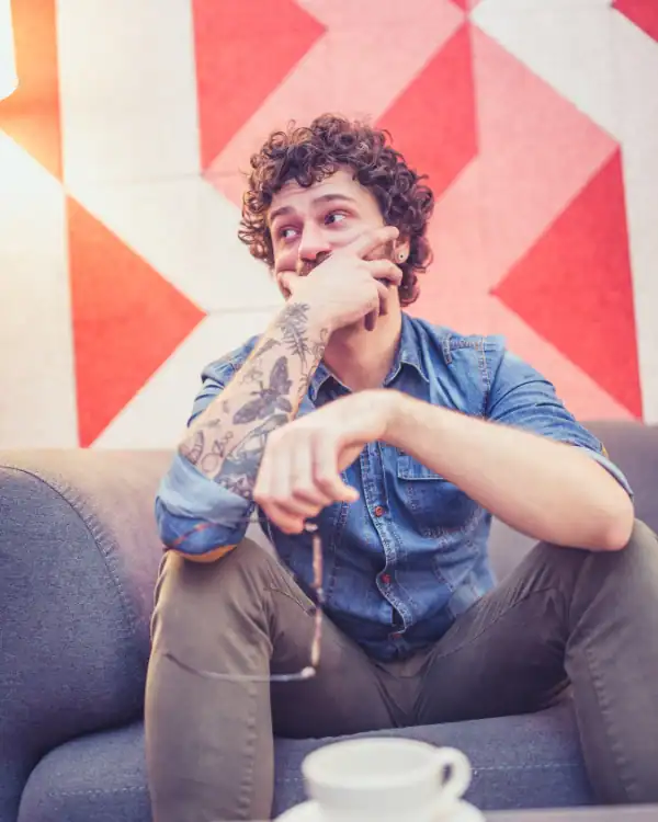 A man with curly hair and tattoos sits on a couch, resting his hand on his chin and holding glasses—perhaps reflecting during an adhd counseling session. He wears a denim shirt as a cup sits on the table before a red geometric background.