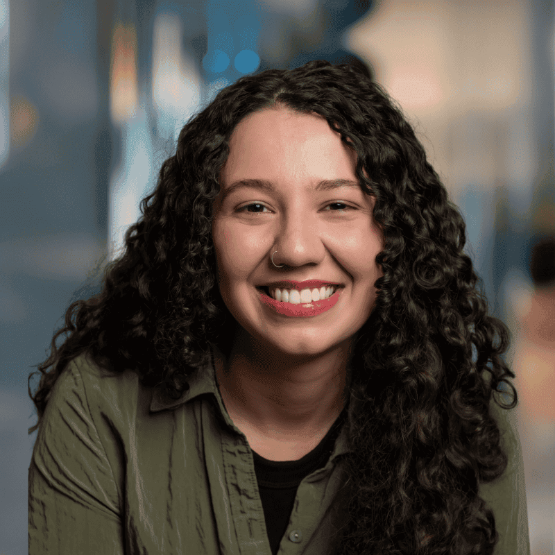A young woman with long, curly dark hair and a nose ring smiles warmly. She is wearing an olive-green shirt and is indoors, with a softly blurred background.