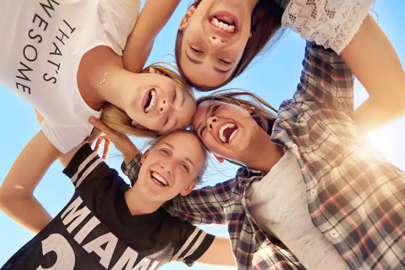 Four smiling teenage girls stand in a circle with their heads together, looking down at the camera against a blue sky, laughing and enjoying each others company.