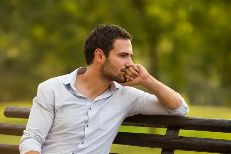 A man with short dark hair and a beard sits on a wooden bench outdoors, resting his chin on his hand and looking thoughtfully into the distance. The background is green and blurred, suggesting a park or garden setting.