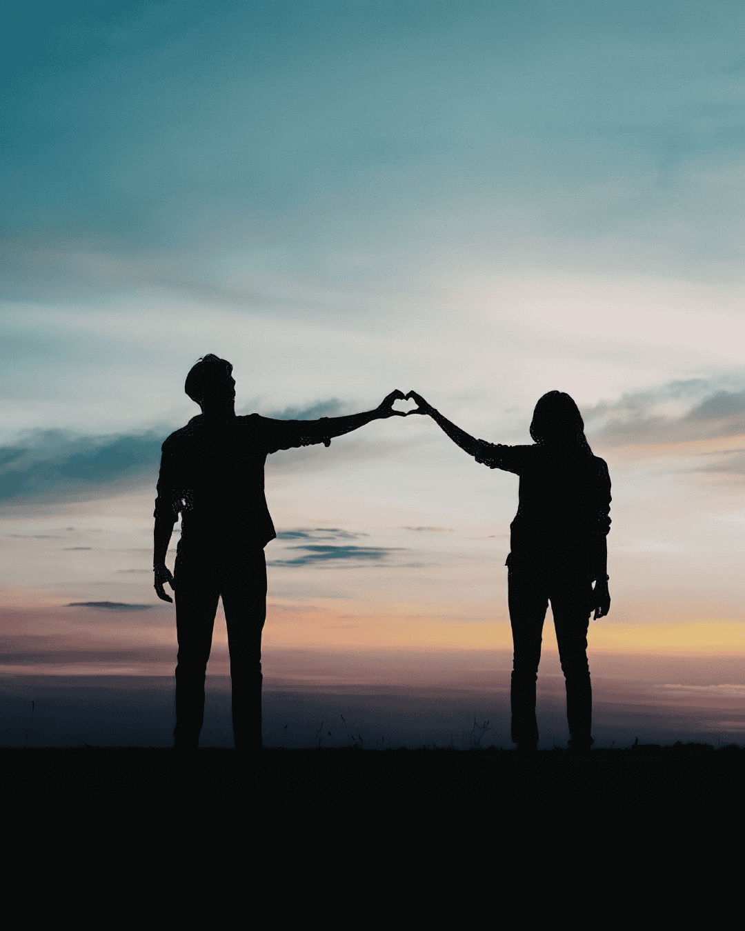 Silhouettes of two people standing apart at sunset, reaching out and touching hands to form the shape of a heart against a colorful sky.