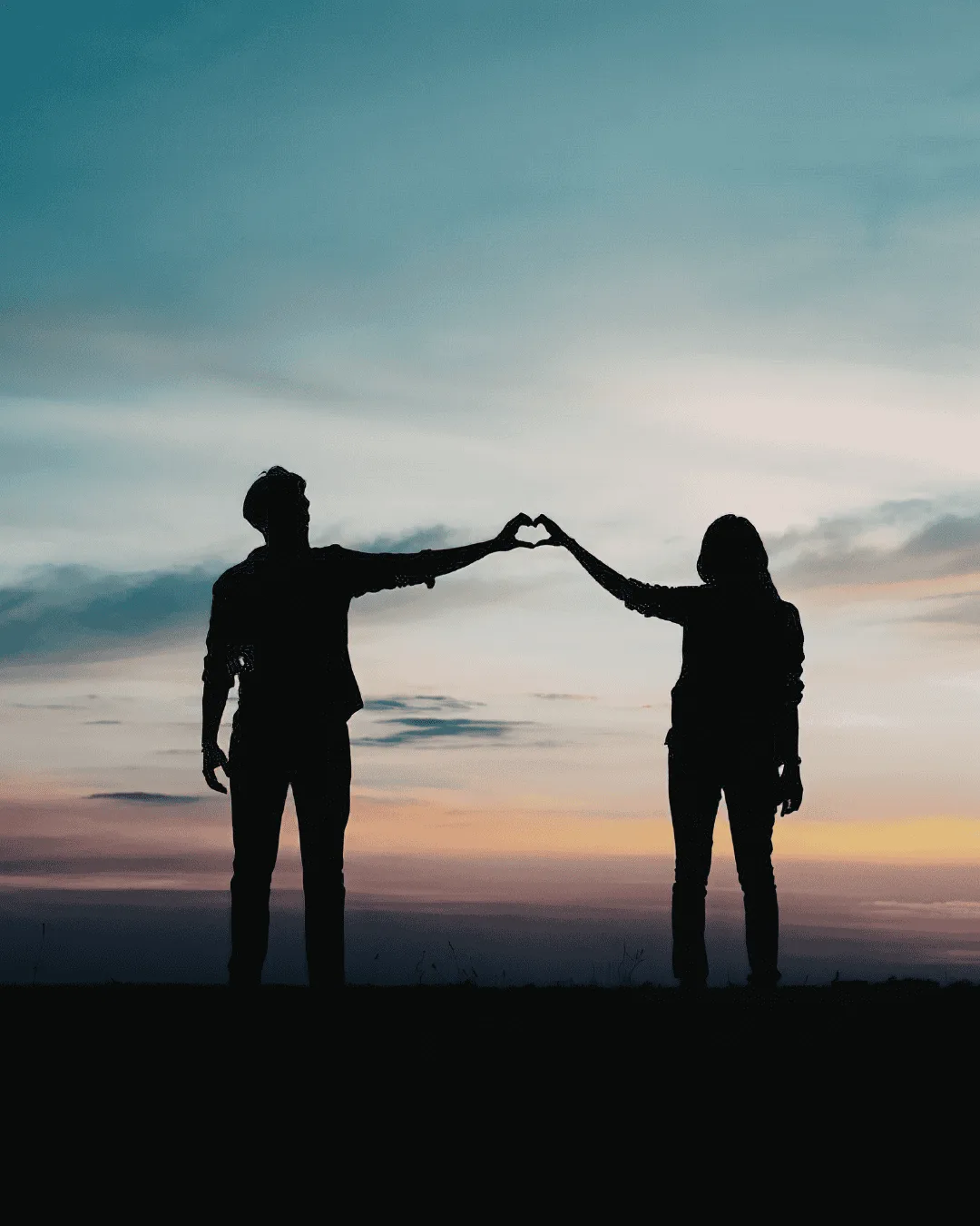 Silhouettes of two people standing apart at sunset, reaching out and touching hands to form the shape of a heart against a colorful sky.