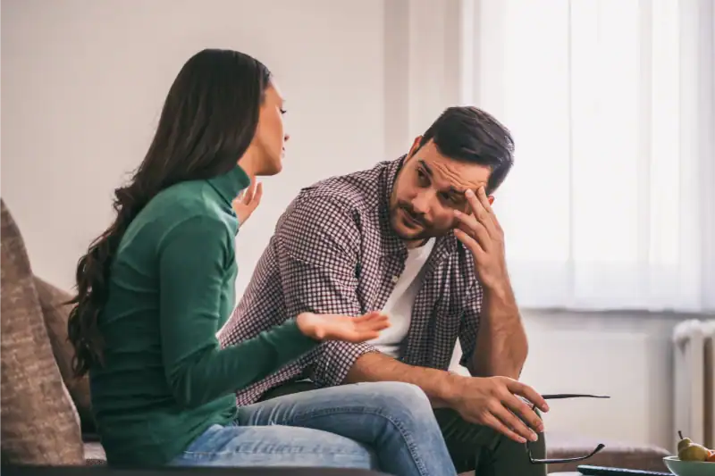 A woman and a man sit on a couch having a serious conversation. The woman gestures with her hands while the man listens with a concerned expression, resting his head on his hand.