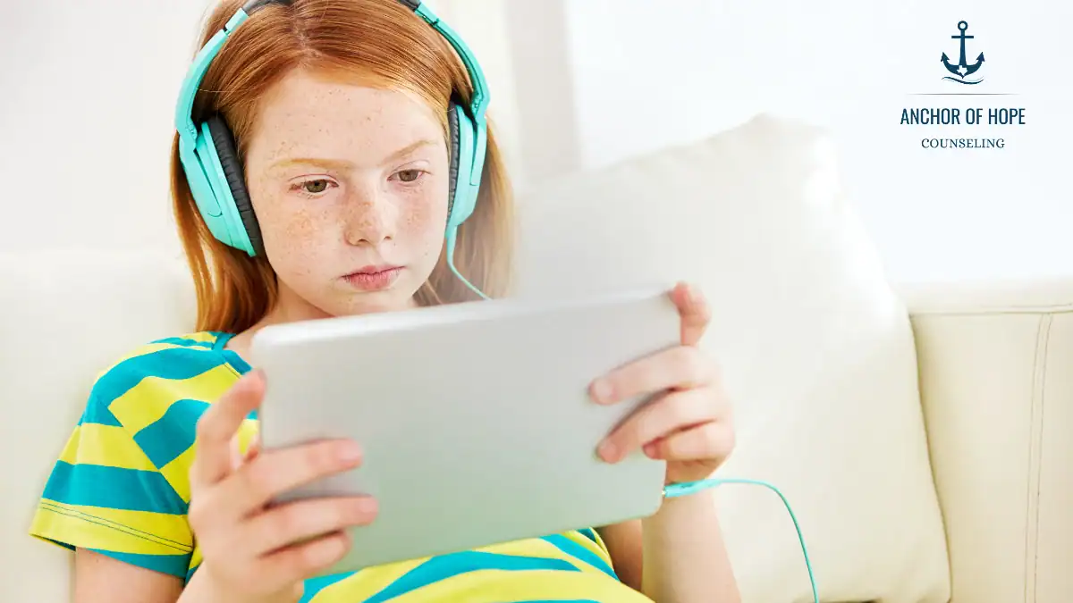 A young girl with red hair wearing blue headphones sits on a white couch, focused on a tablet. She wears a yellow and green striped shirt. The Anchor of Hope Counseling logo is visible in the top right corner.