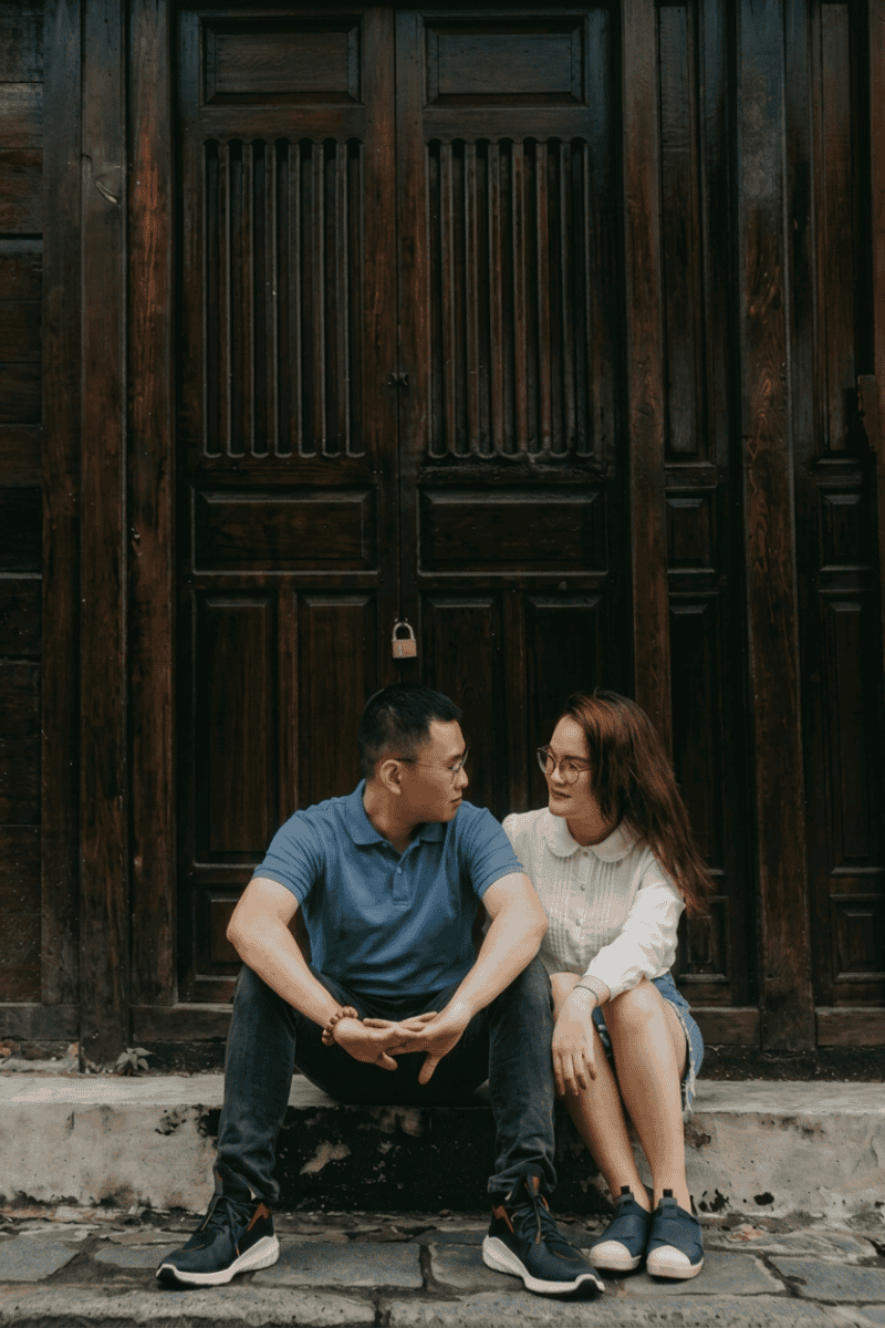 A man and woman sit close together on steps in front of a large wooden door, looking at each other and smiling. The man wears a blue shirt; the woman wears glasses and a white top.