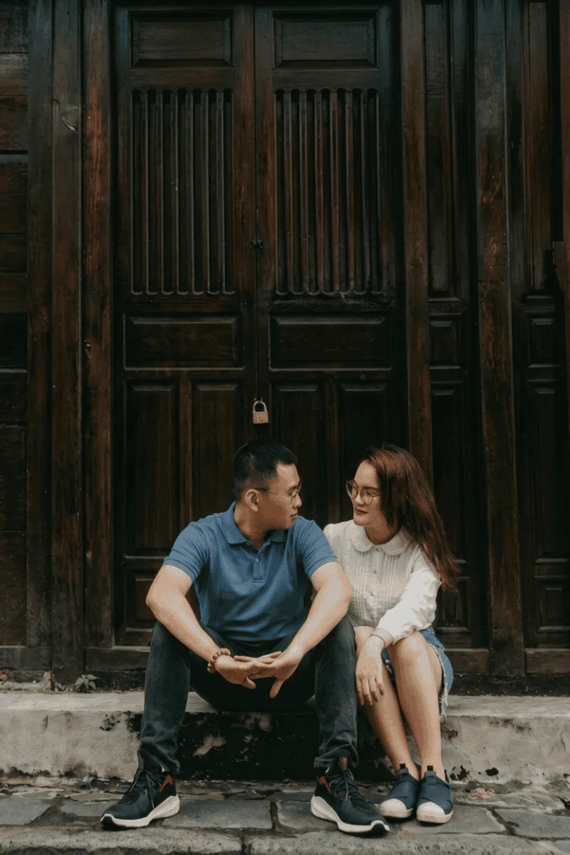 A man and woman sit close together on steps in front of a large wooden door, looking at each other and smiling. The man wears a blue shirt; the woman wears glasses and a white top.