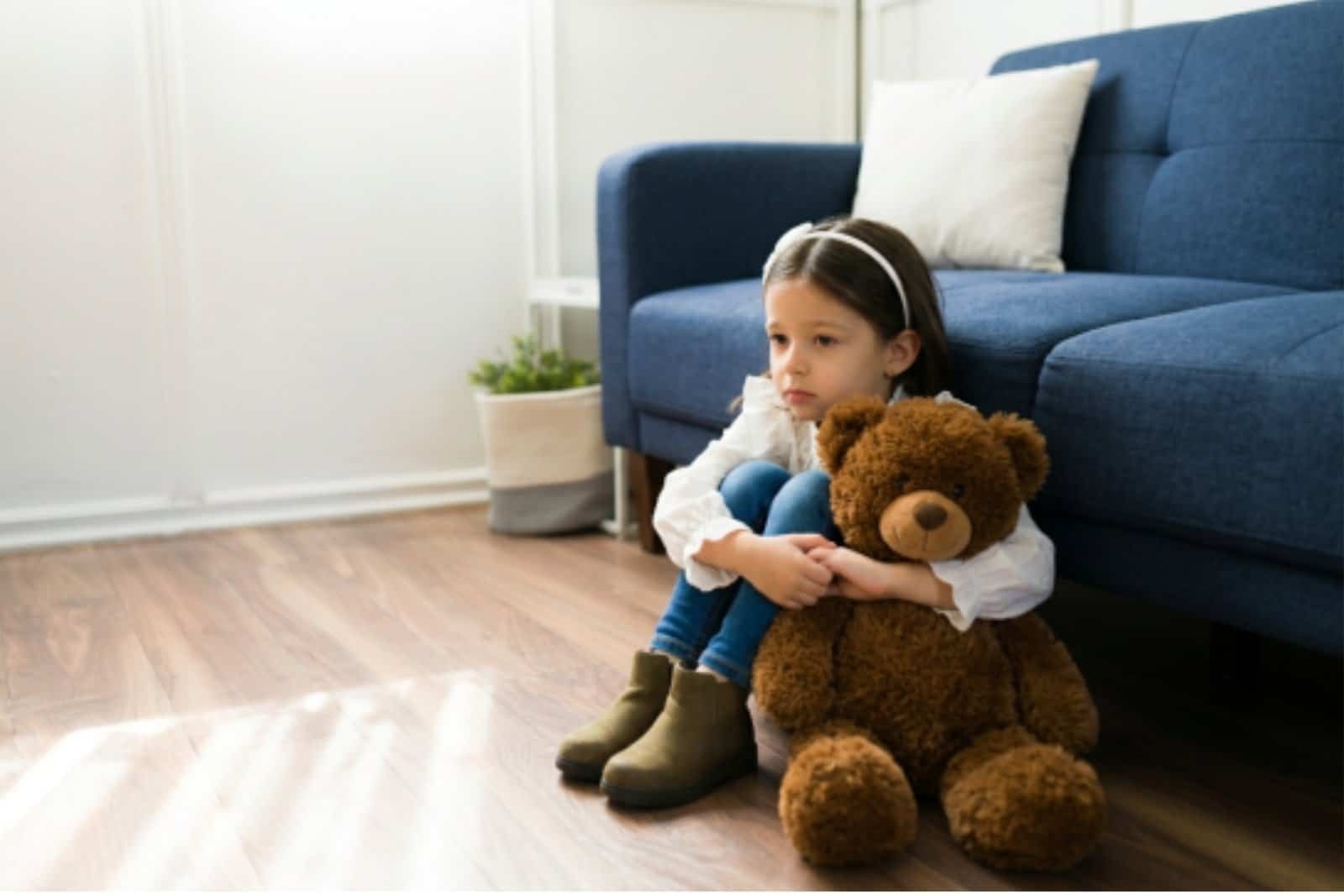 A young girl sits on a wooden floor next to a blue sofa, hugging a large brown teddy bear and looking thoughtful. There is a potted plant in the background.