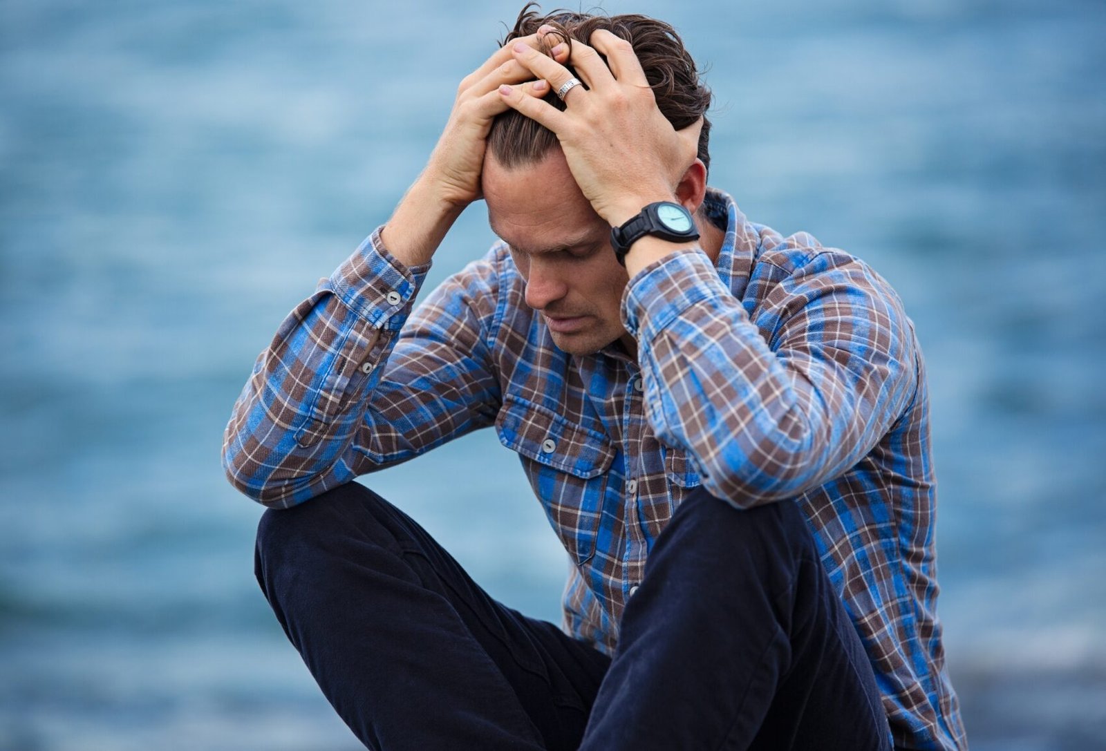 A man in a plaid shirt sits by the water, holding his head with both hands and looking down, appearing stressed or deep in thought.