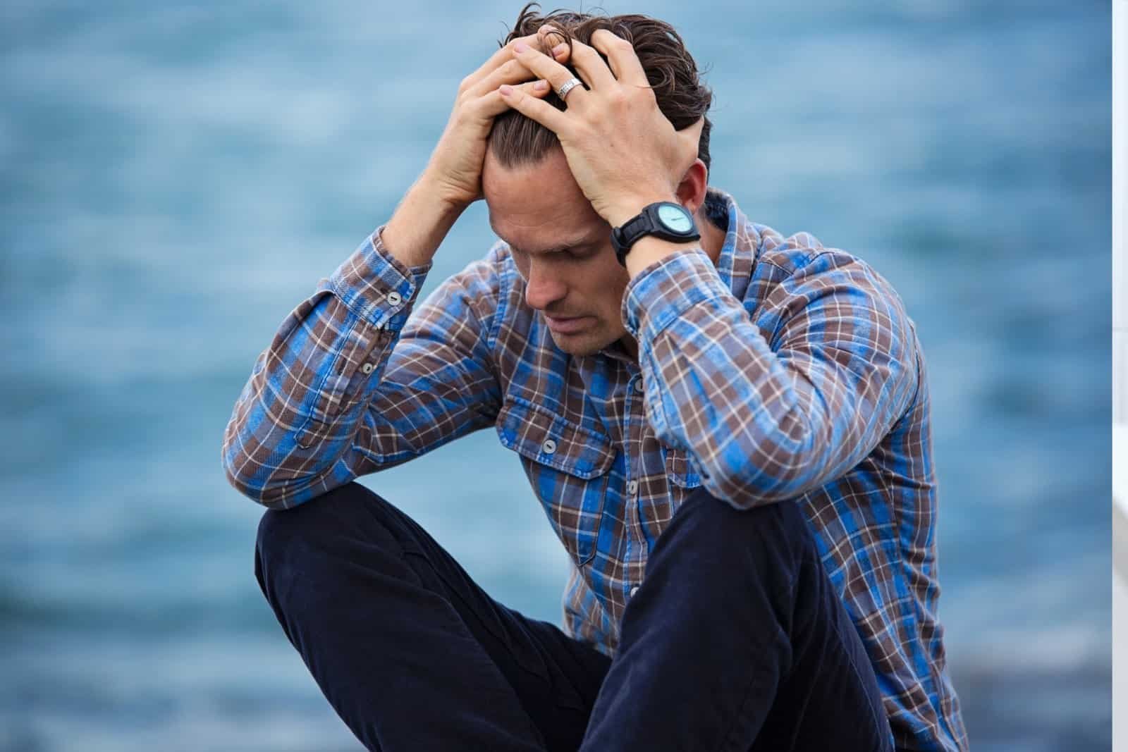 A man in a plaid shirt sits by the water, holding his head with both hands and looking down, appearing stressed or deep in thought.